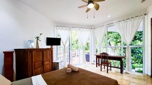 A bedroom with a bed, a desk, and a chair. There is a television on the wall and a ceiling fan.