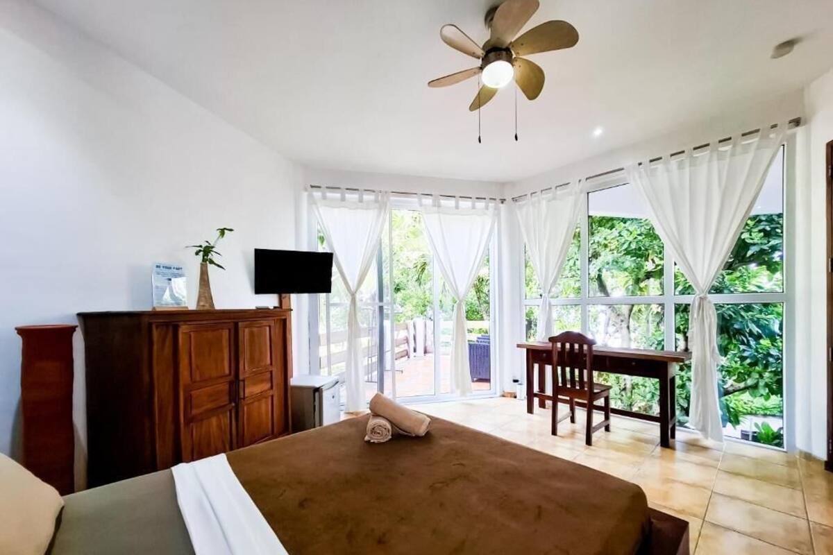 A bedroom with a bed, a desk, and a chair. There is a television on the wall and a ceiling fan.