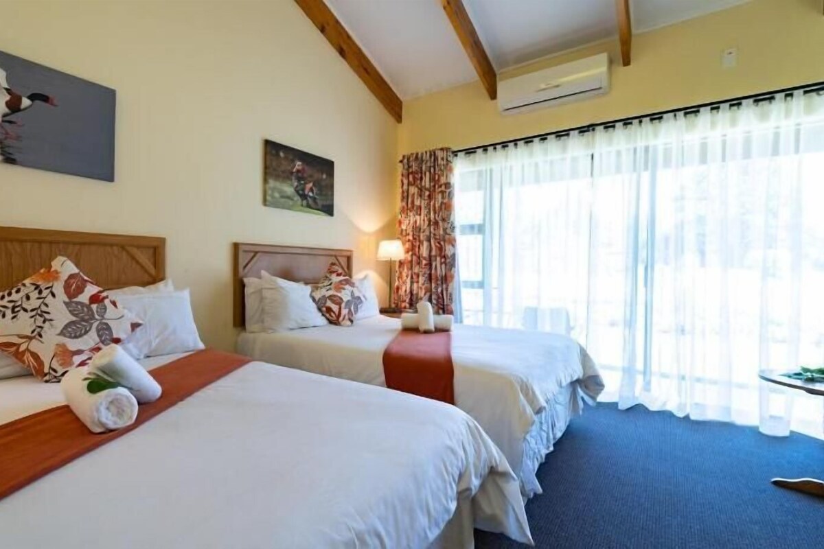 Une chambre d’hôtel avec deux lits, une tête de lit en bois, un tableau, une fenêtre avec des rideaux et un appareil de climatisation.