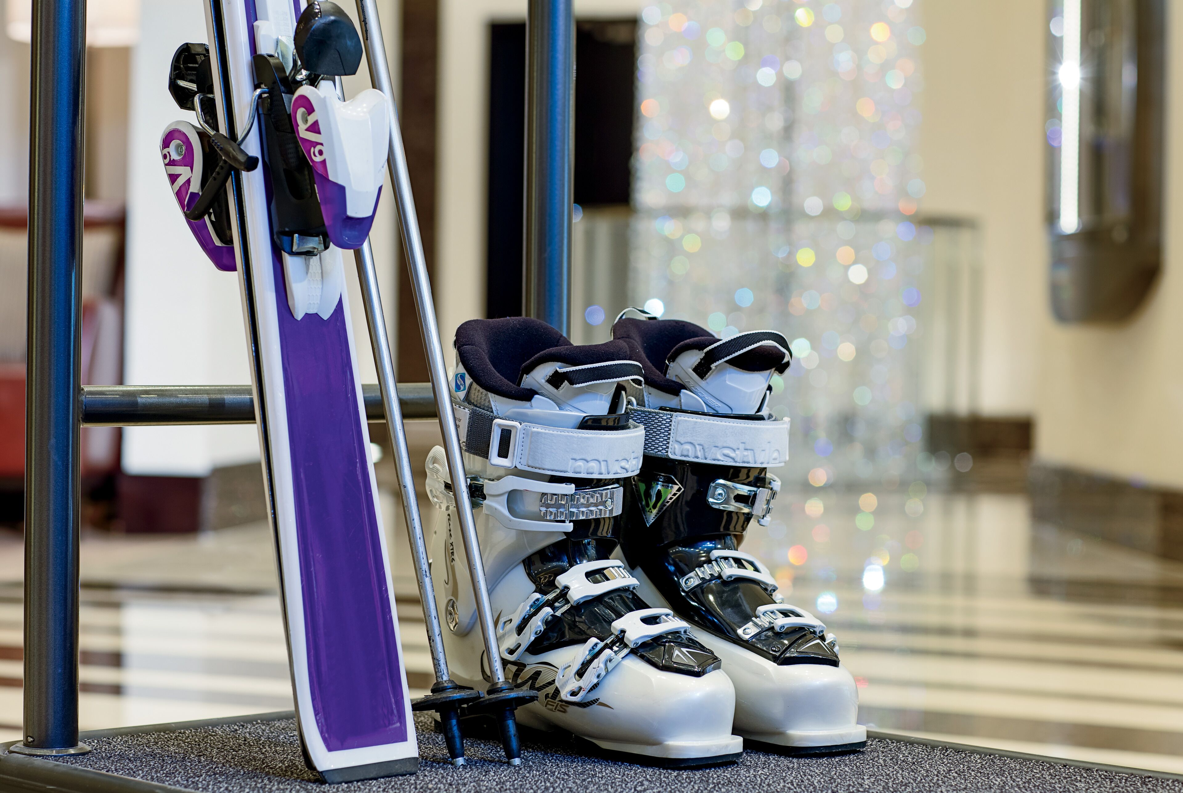 A pair of skis and ski boots leaning against a metal pole in a well-lit indoor area with a reflective floor and decorative lights in the background.
