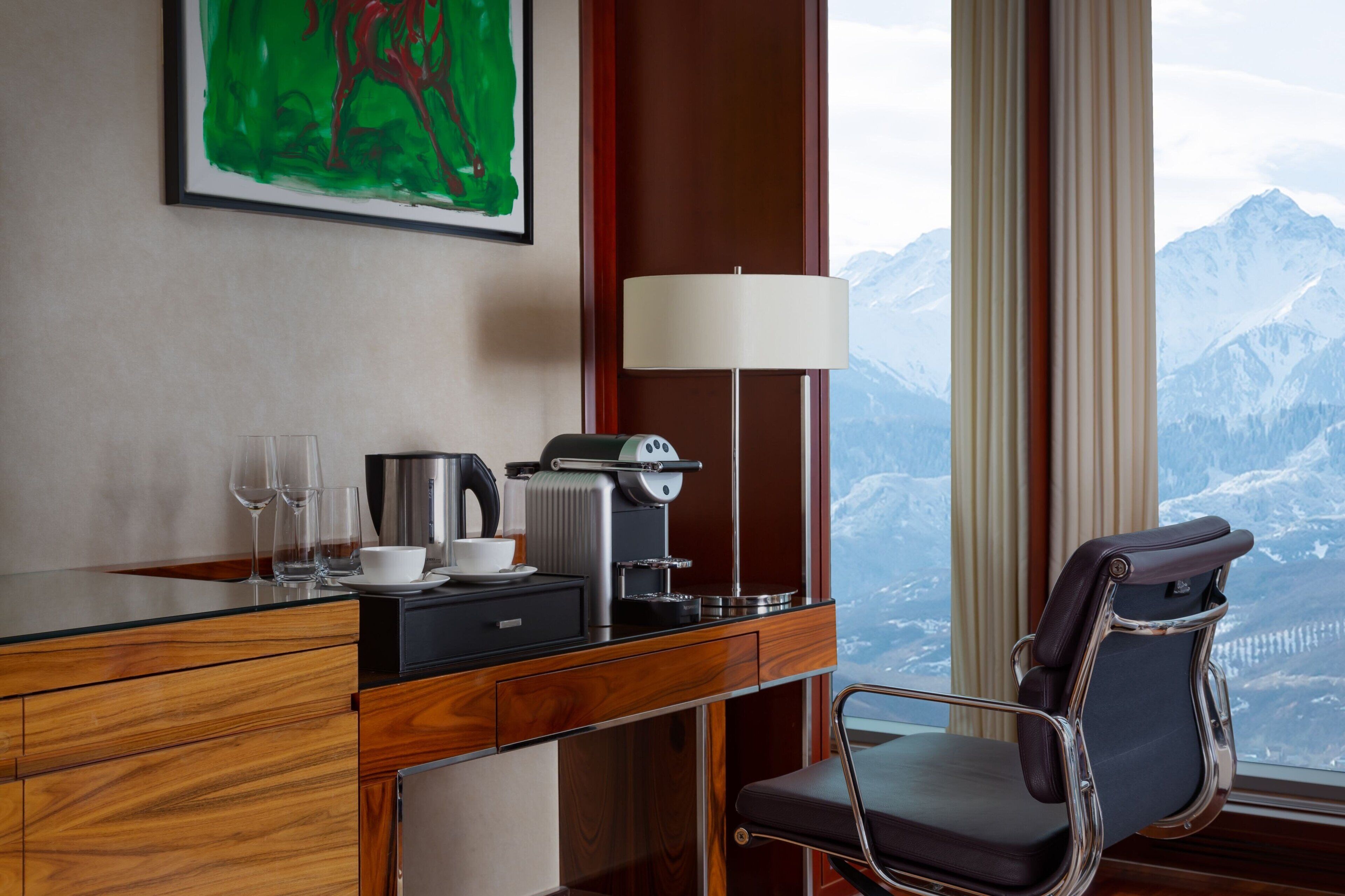 A hotel room with a wooden desk, a coffee machine, a chair, and a view of mountains.