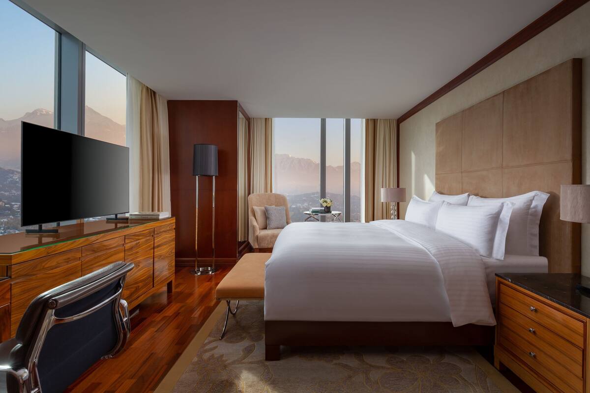 A hotel room with a large bed, a TV on a wooden dresser, a chair, and a view of mountains through large windows.