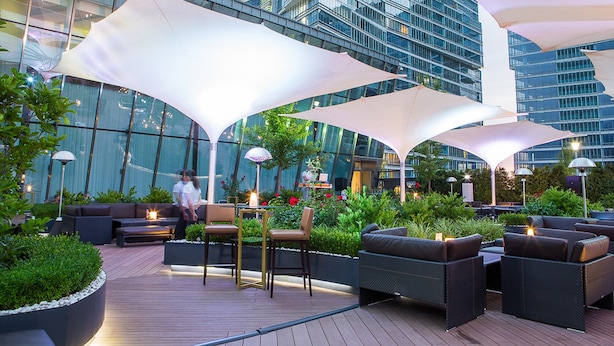 An outdoor seating area with modern furniture and large umbrellas, surrounded by lush greenery and a glass building in the background. - The Ritz-Carlton, Almaty (Almaty)