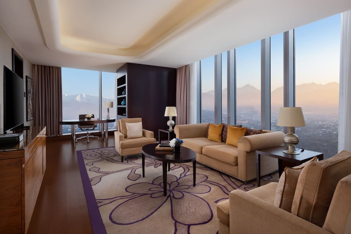 A modern hotel room with a large window offering a mountain view, a sofa, a coffee table, and a TV.