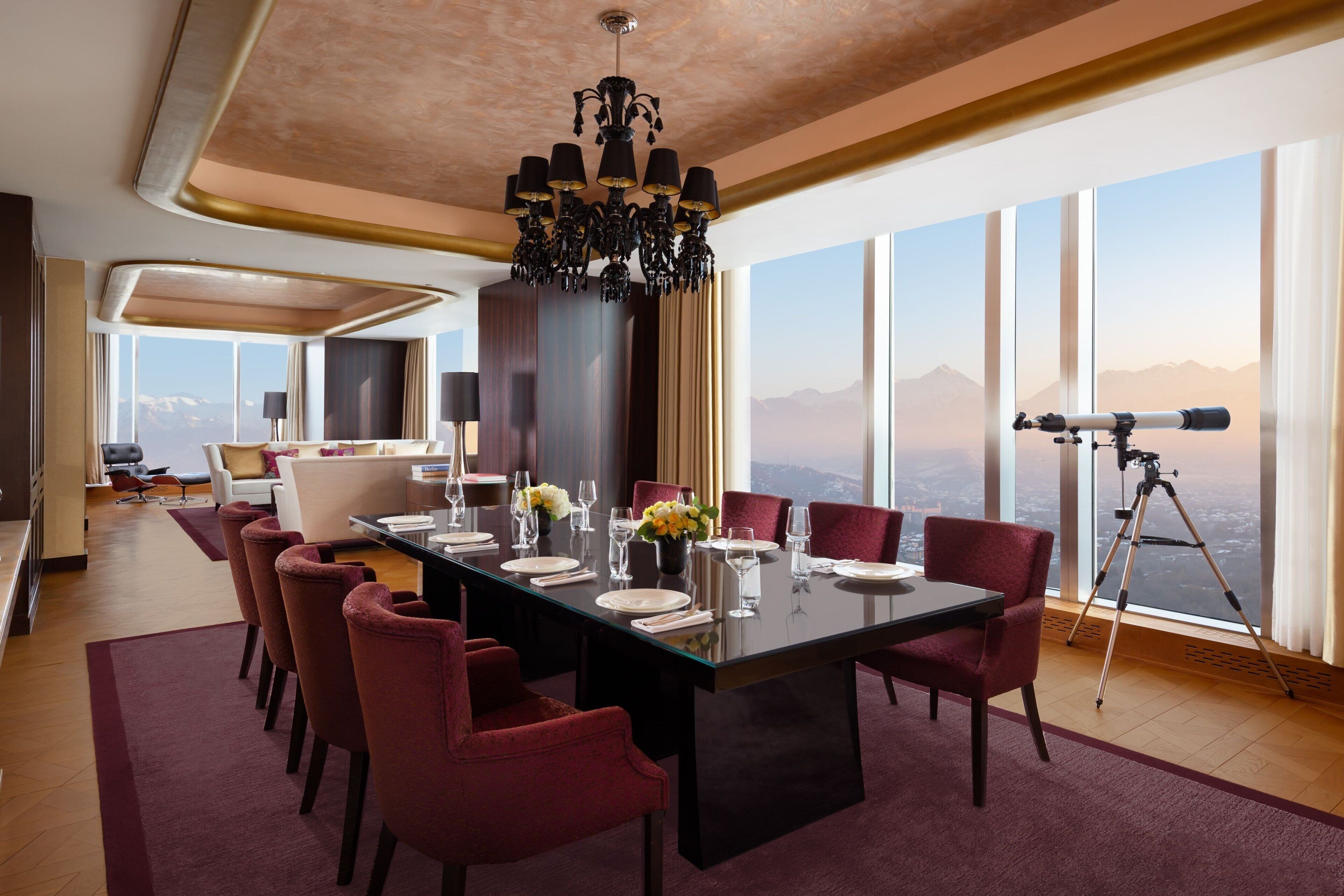A dining area with a large table set for six, red chairs, a chandelier, and a telescope by the window.