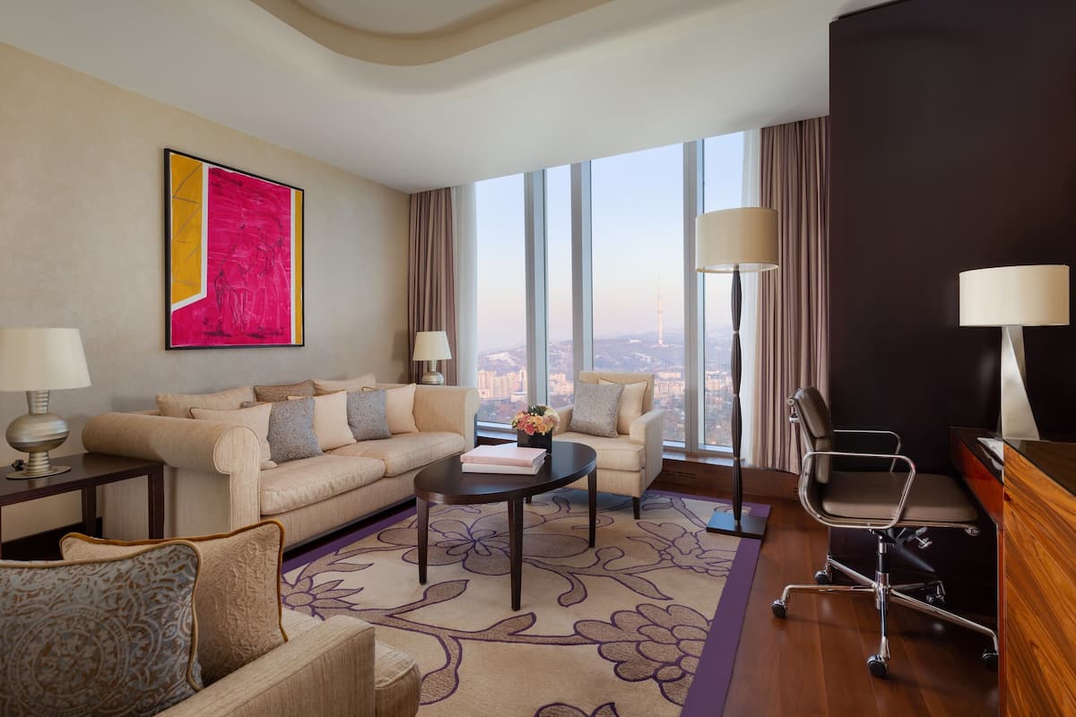 A modern hotel room with a large window offering a city view, a beige sofa set, a round coffee table, and a desk with a chair.