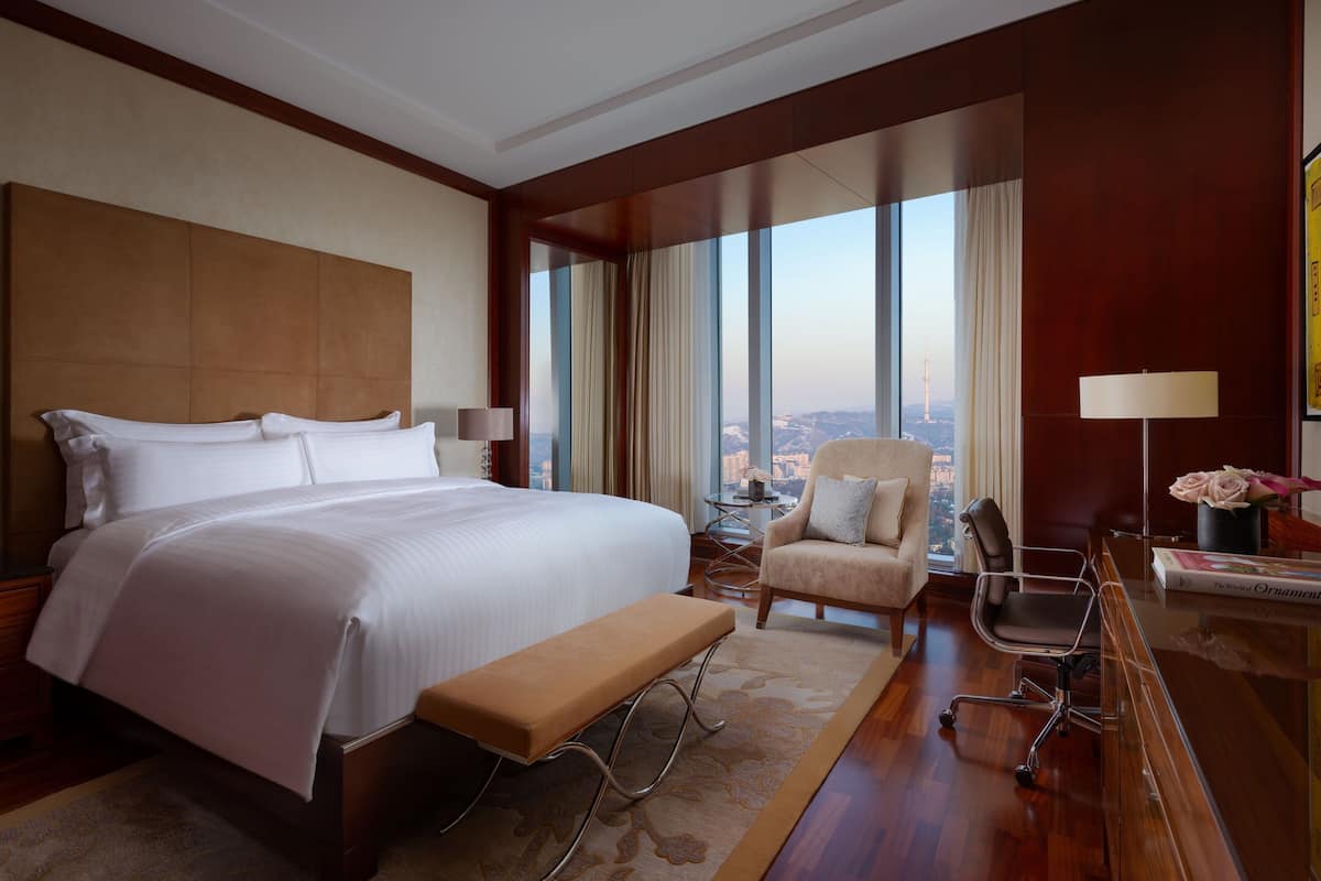 A hotel room with a large bed, a desk with a lamp, a chair, and a view of the city.