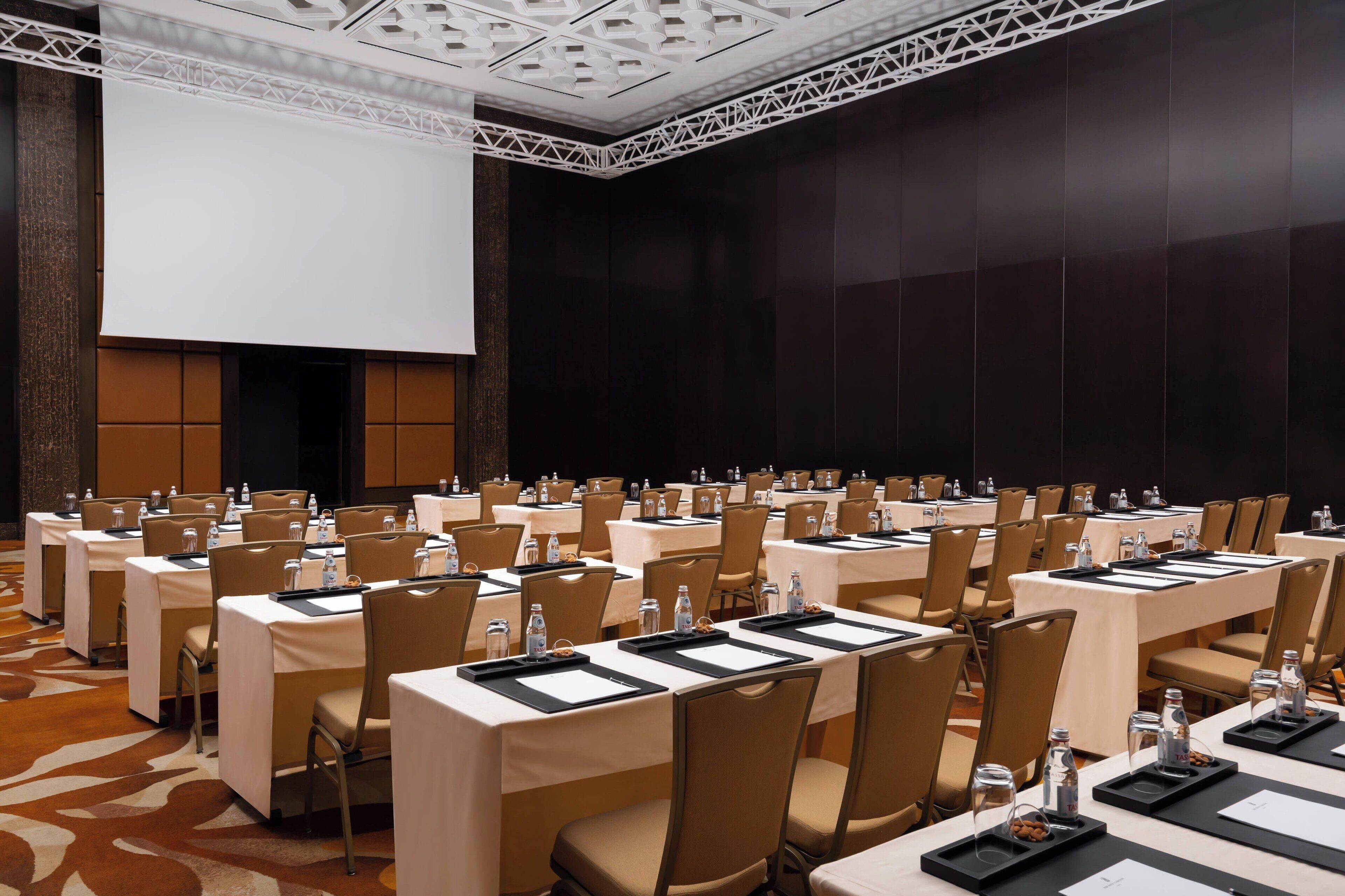 A conference room with rows of tables, chairs, and a large screen.