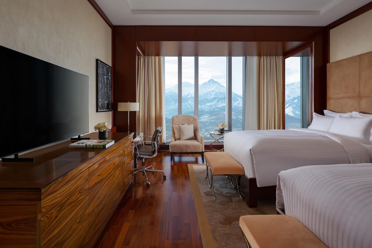 A hotel room with a large bed, a desk with a TV, a chair, and a view of mountains through large windows.