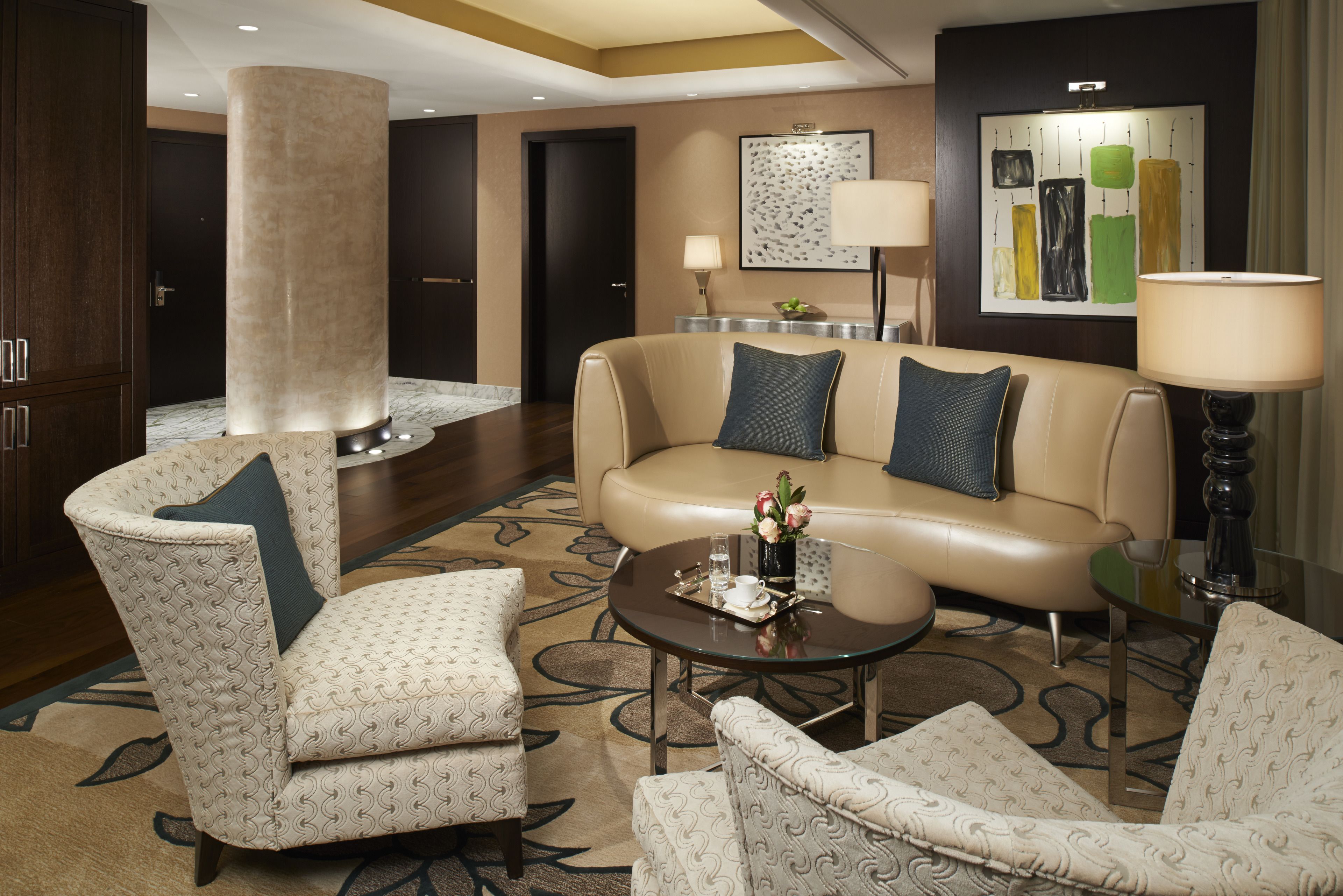 A modern hotel lobby with a beige sofa, patterned armchairs, a round coffee table, and a large pillar.