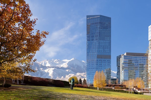 A modern building with a glass facade, a mountain backdrop, and a park with autumn trees.