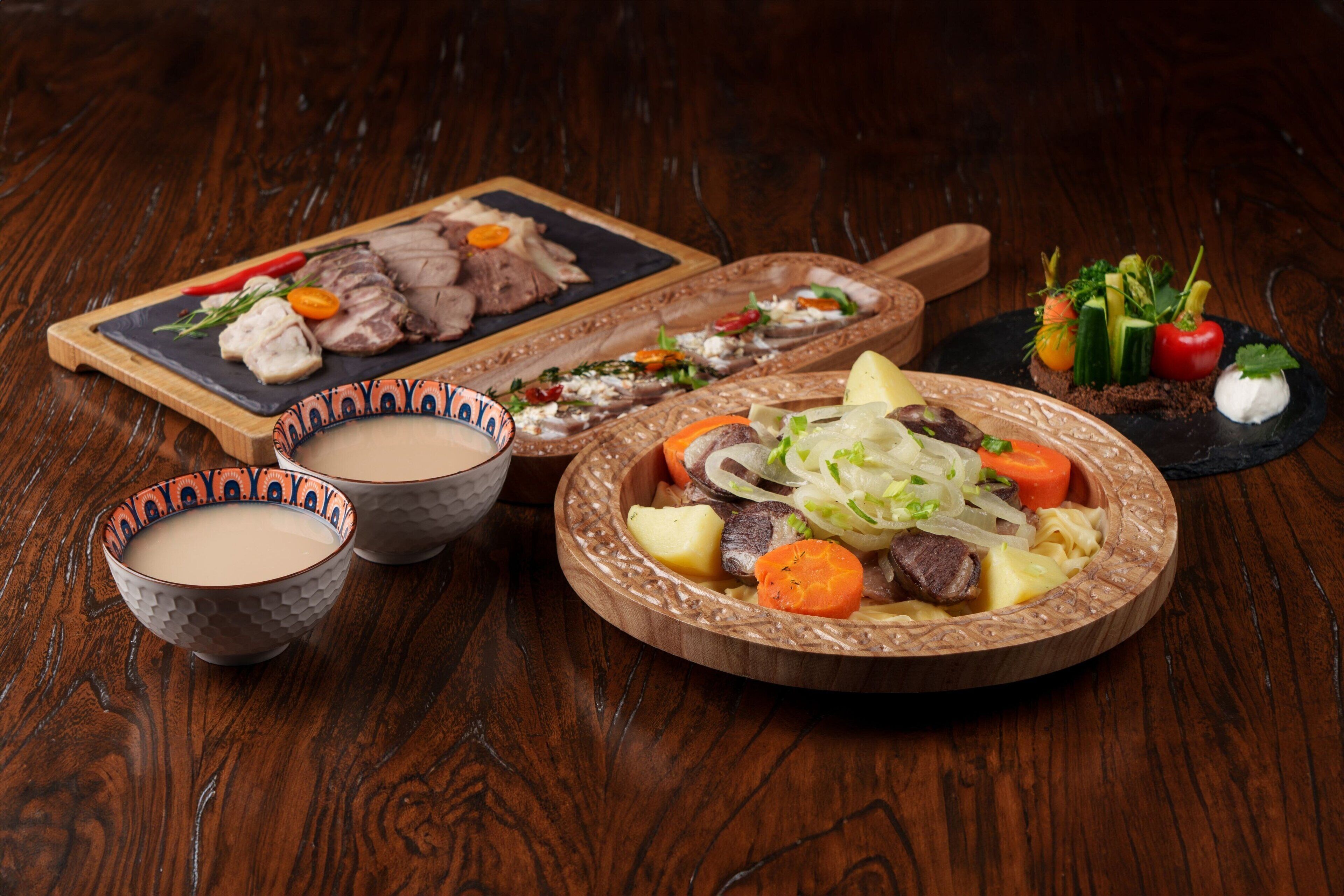 A wooden table with a variety of dishes including a bowl of soup, a plate of sliced meat, and a bowl of salad.