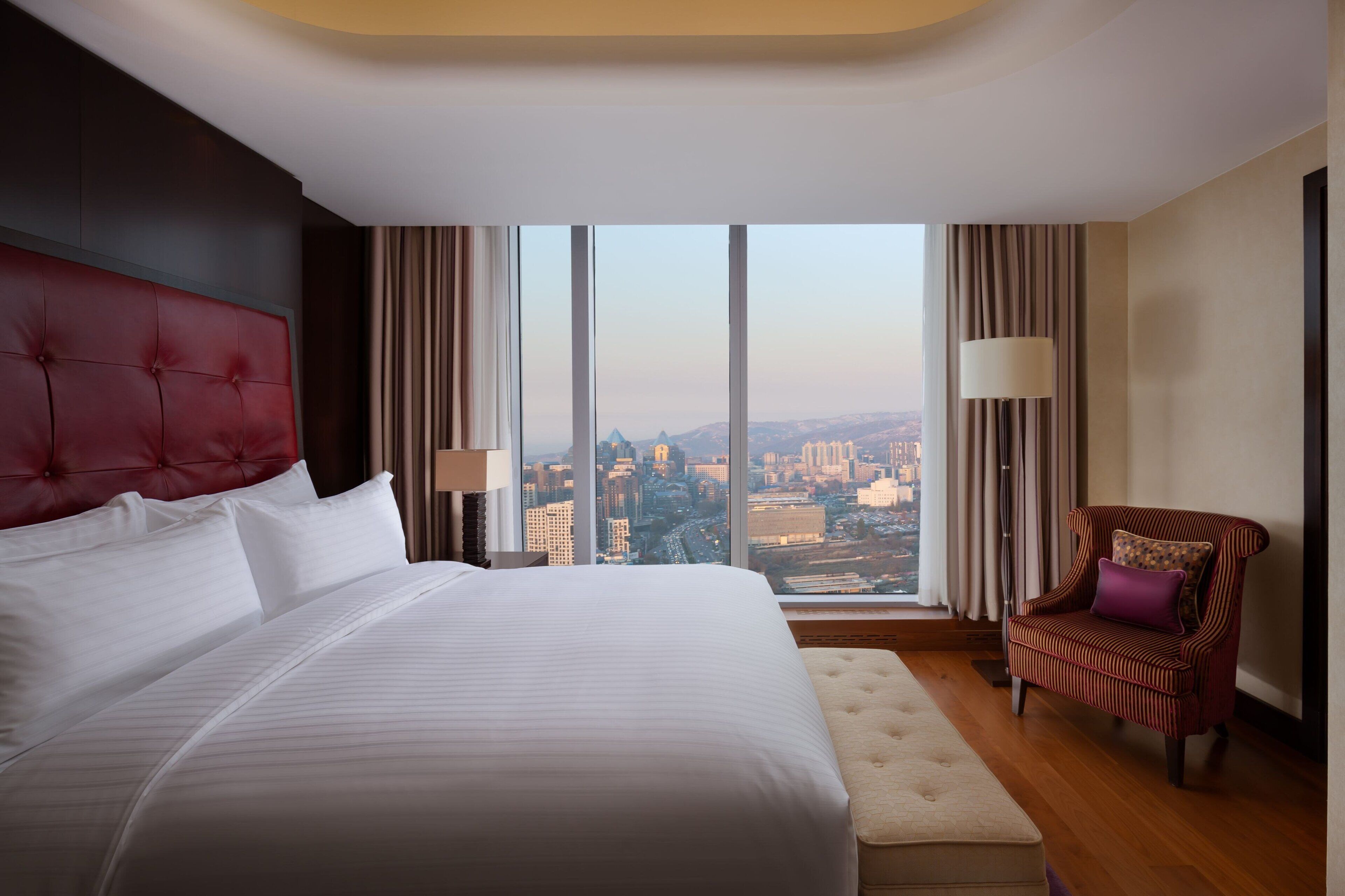 A hotel room with a large bed, a red headboard, a view of a cityscape, a chair with a cushion, and a lamp.