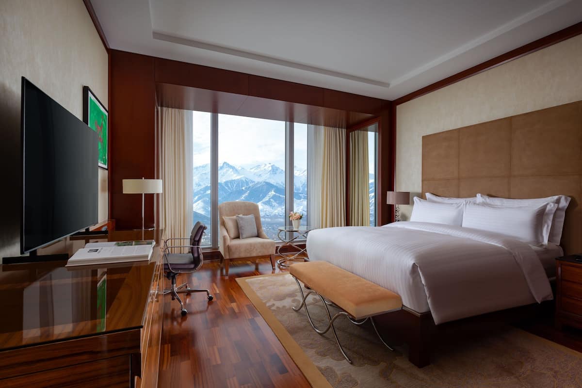 A hotel room with a large bed, a desk with a flat-screen TV, a chair, and a view of mountains through large windows.