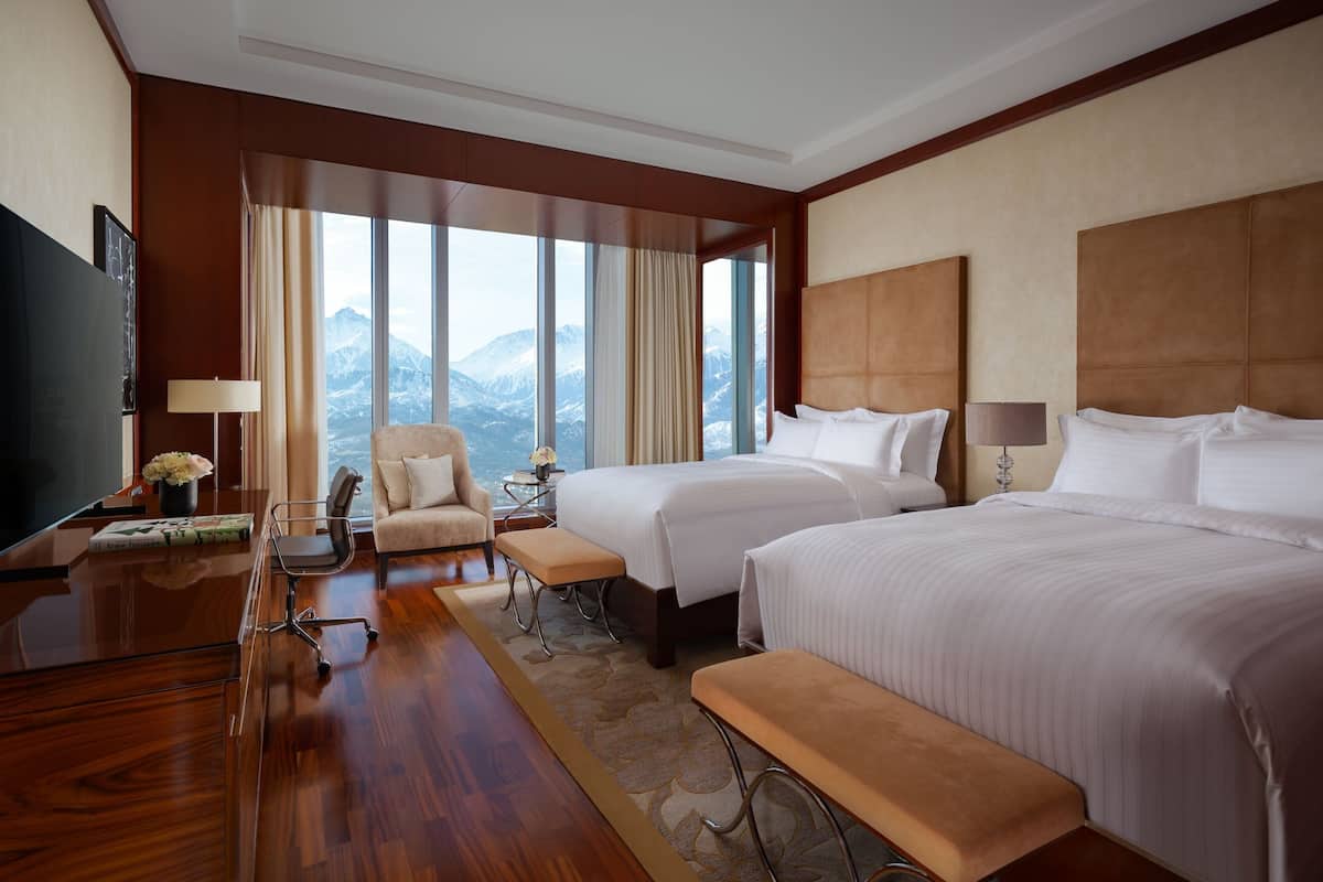 A hotel room with two beds, a desk, a chair, and a TV.