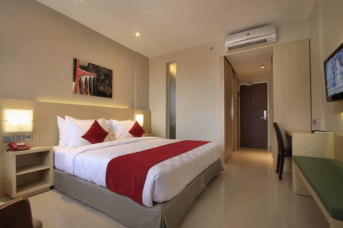 A hotel room with a bed, bedside tables, a desk, a chair, a TV, and an air conditioning unit.