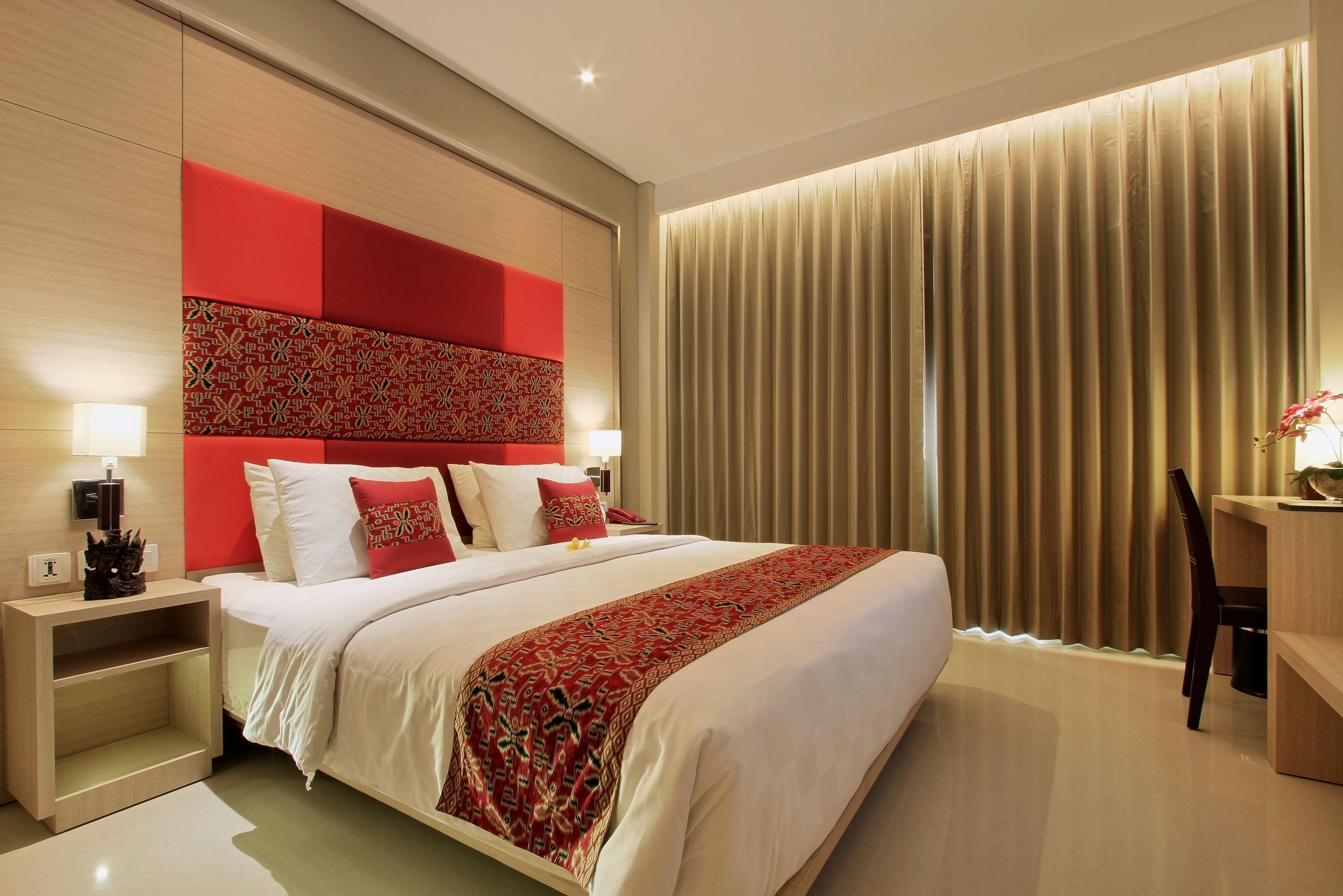 A modern hotel room with a large bed, red accent wall, and a desk with a chair.