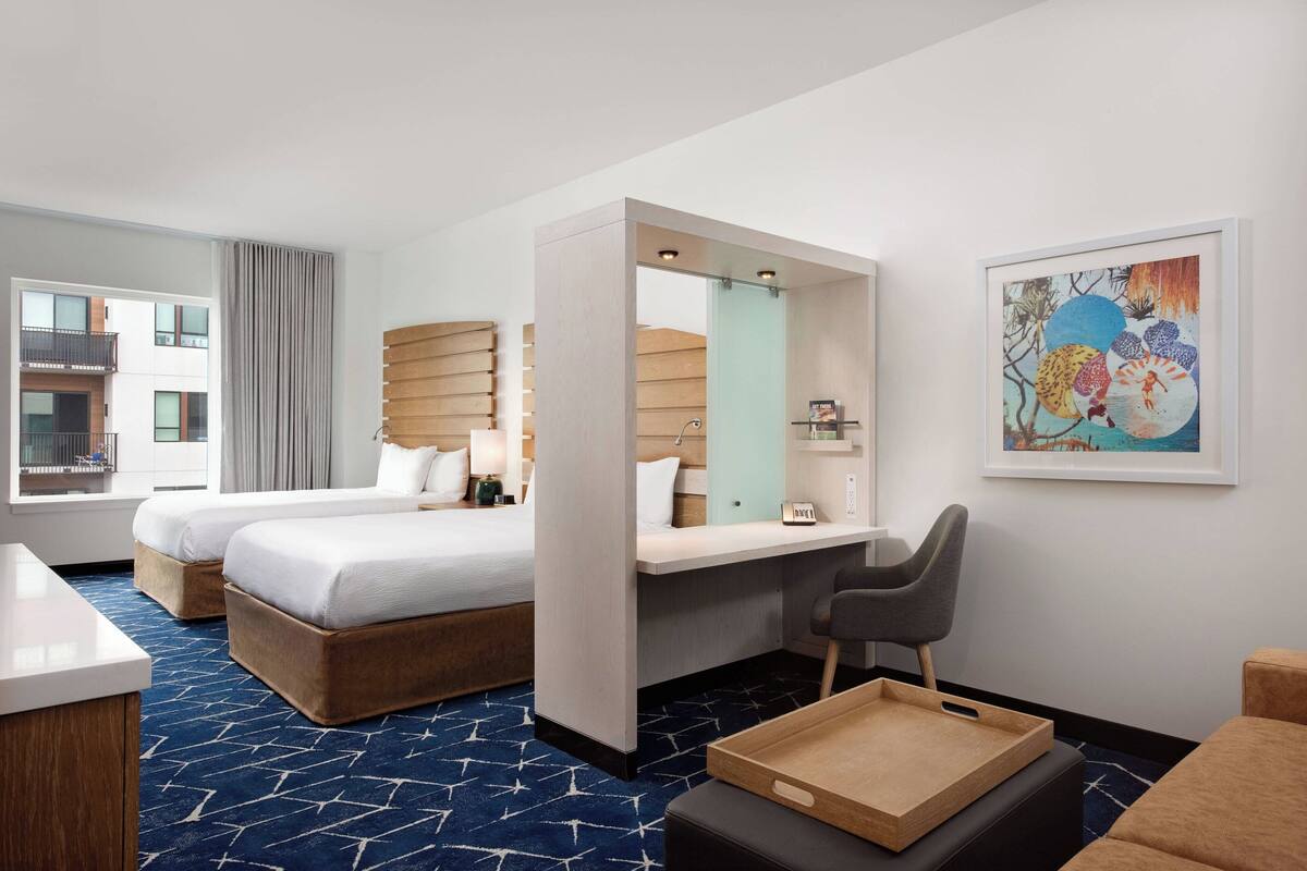 Ein Hotelzimmer mit zwei Betten, einem Schreibtisch, einem Stuhl, einem Tablettisch und einem großen Fenster.
