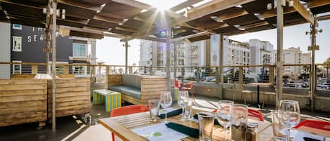 An outdoor seating area with wooden tables and chairs, a view of the cityscape, and a pergola structure.