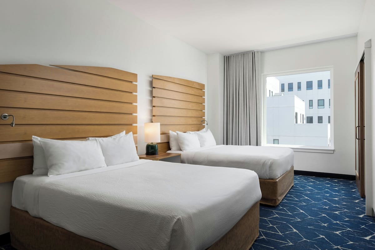 Ein Hotelzimmer mit zwei Betten, Holz-Kopfteilen, einem großen Fenster mit Vorhängen und einem gemusterten Teppich.