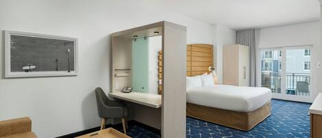 A hotel room with a bed, a desk, a chair, a small table, a mirror, and a window with a view of buildings.