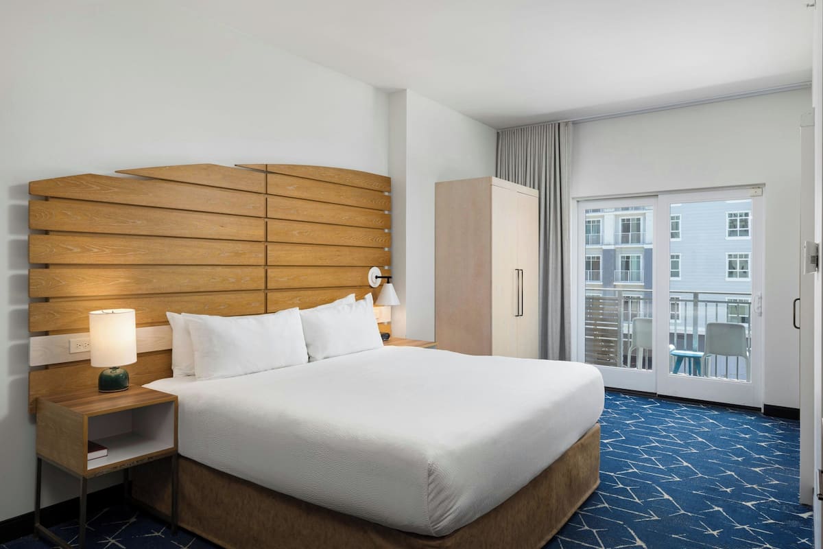 Ein Hotelzimmer mit einem großen Bett, einem hölzernen Kopfteil, einem Nachttisch mit Lampe, einem Kleiderschrank und einem Balkon mit Blick auf Gebäude.