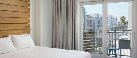 A hotel room with a large bed, a view of the city through a sliding glass door, and a balcony with chairs.