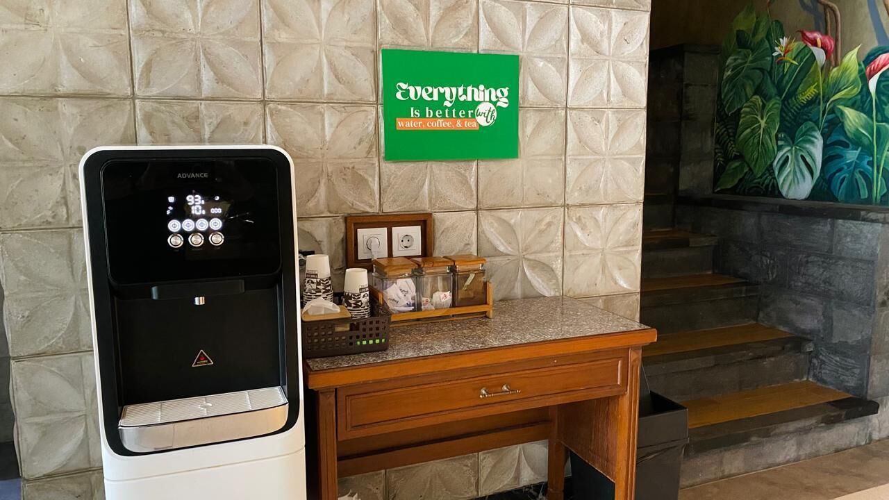 A water dispenser, a coffee machine, a small table with a tray of coffee cups, a framed picture, and a green sign on the wall.