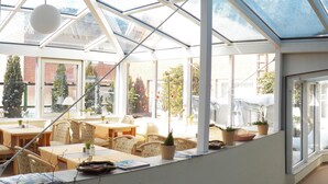 Una habitación iluminada por el sol con techo de vidrio, amueblada con mesas y sillas, y plantas en macetas.