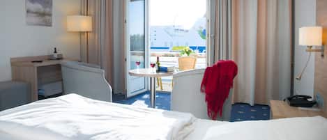 A hotel room with a bed, a desk, a chair, a table with a bottle and glass, and a view of the sea and a ship.