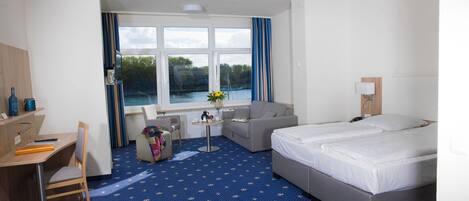 A hotel room with a large bed, a desk, a chair, a sofa, and a view of a lake through the windows.