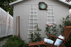 A wooden shed with a white exterior, a round window, and a white ladder. There is a wooden chair with a cushion and a plant with pink flowers.