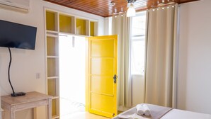 Habitación de hotel con una cama, un televisor sobre una mesita, una puerta amarilla y una ventana con cortinas.