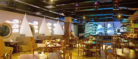 A restaurant with wooden tables and chairs, a stage with a large screen, and a view of buildings in the background.