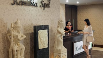 Une salle d’attente avec un comptoir en marbre, une réceptionniste et deux clients. On y trouve des statues décoratives et une enseigne indiquant « Karibia Spa ».