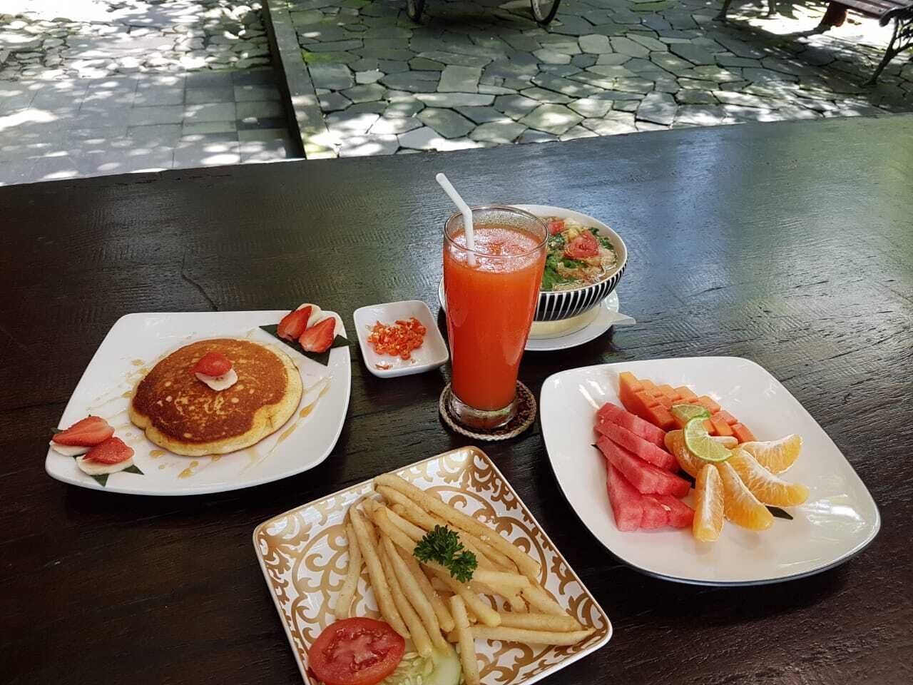 Ein Tisch mit Frühstücksangeboten, darunter Pfannkuchen, ein Glas Tomatensaft, eine Salatschüssel, eine Schale mit Wassermelone und Orangen sowie eine Portion Pommes.