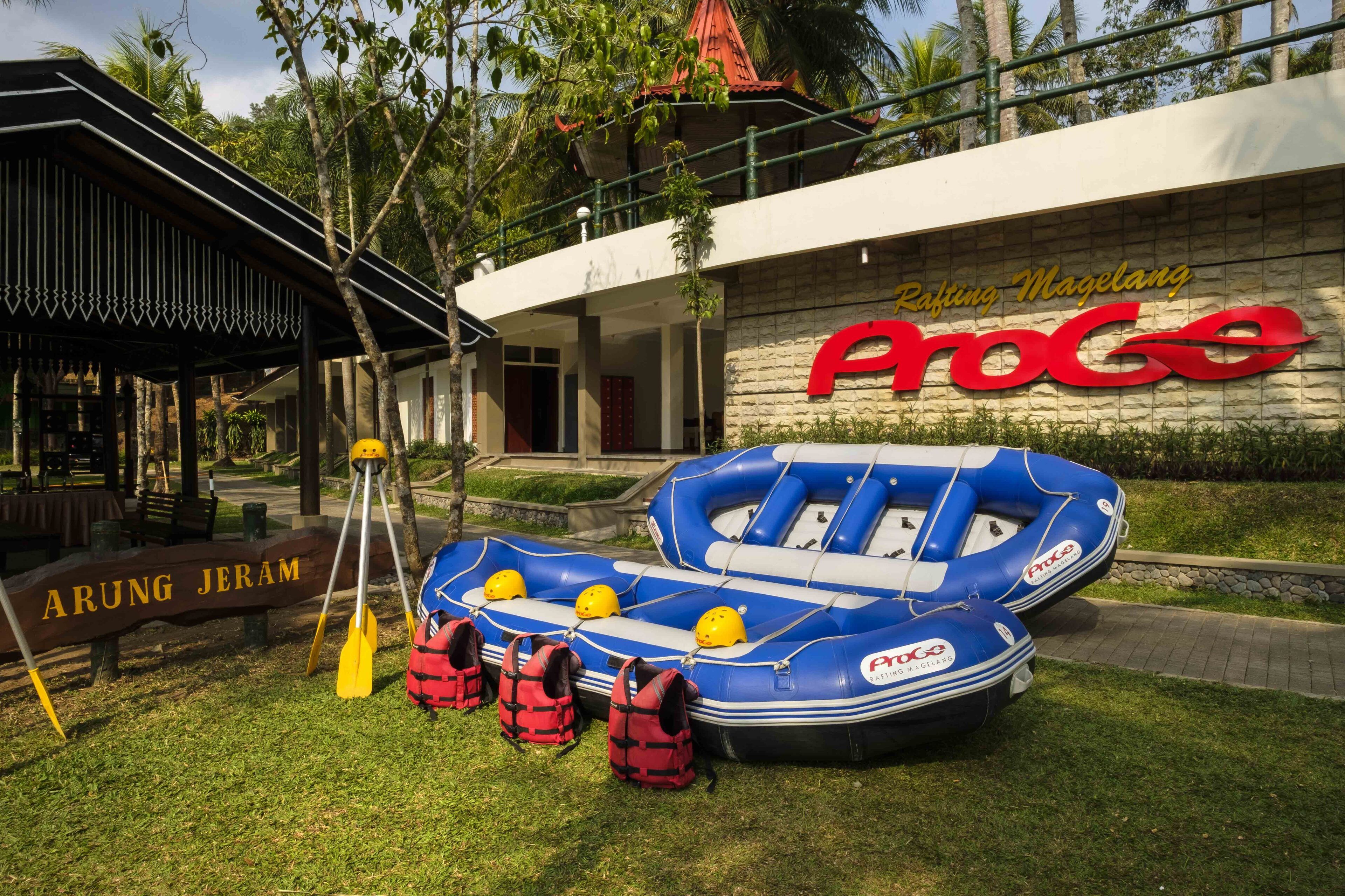 A blue inflatable raft with life jackets and paddles, a sign reading "ARUNG JERAM," and a building with "Rafting Magelang ProGo" signage.