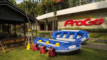 A blue inflatable raft with life jackets and paddles, a sign reading "ARUNG JERAM," and a building with "Rafting Magelang ProGo" signage.