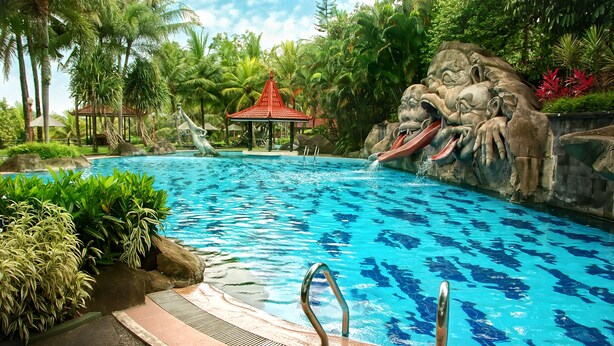 A swimming pool with a waterfall feature, surrounded by tropical vegetation and a gazebo. - Hotel Puri Asri (Magelang)