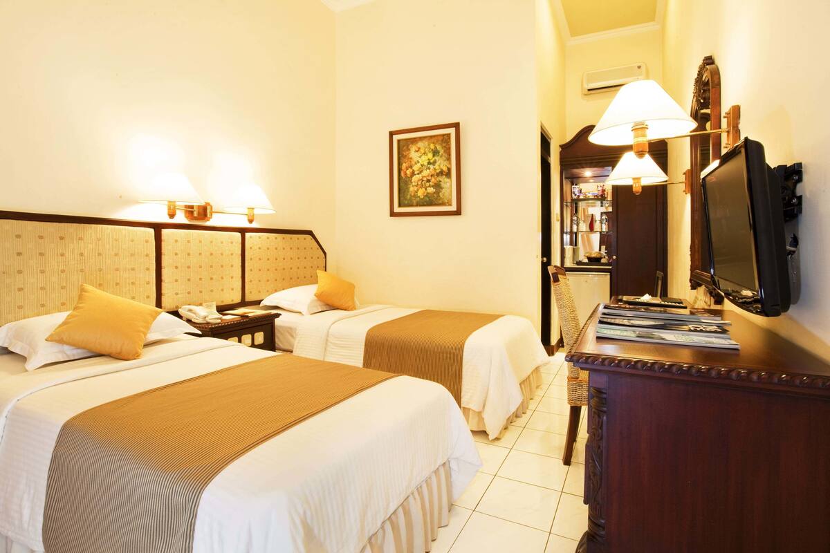 A hotel room with two beds, a desk, a lamp, a television, and a painting on the wall.