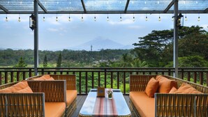 A covered outdoor seating area with wicker furniture and orange cushions, overlooking a mountain and lush greenery. - Hotel Puri Asri (Magelang)
