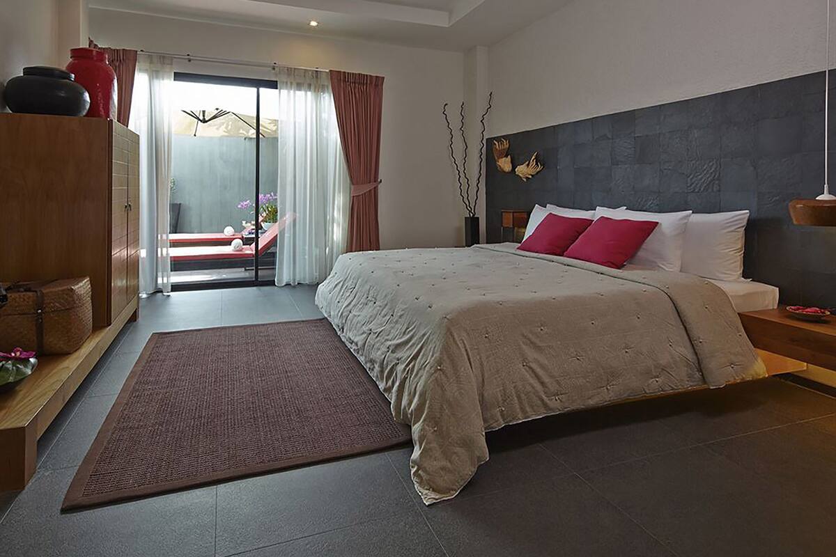 Ein modernes Schlafzimmer mit einem großen Bett, einem Holzschrank, einem Schreibtisch und Blick durch eine Glastür auf einen Außenbereich.