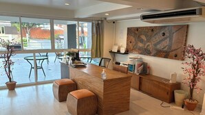 A modern reception area with a wooden reception desk, cushioned seating, a large window, and a wall-mounted air conditioning unit. - Nantra Huahin (Hua Hin)