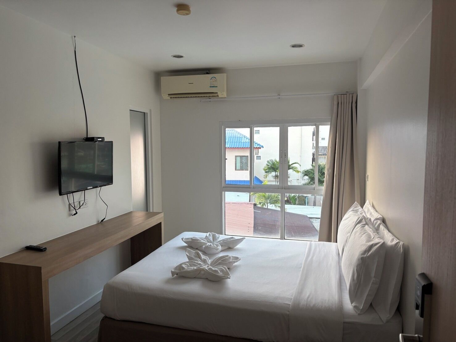 A hotel room with a bed, a TV mounted on the wall, a window with curtains, and an air conditioning unit.