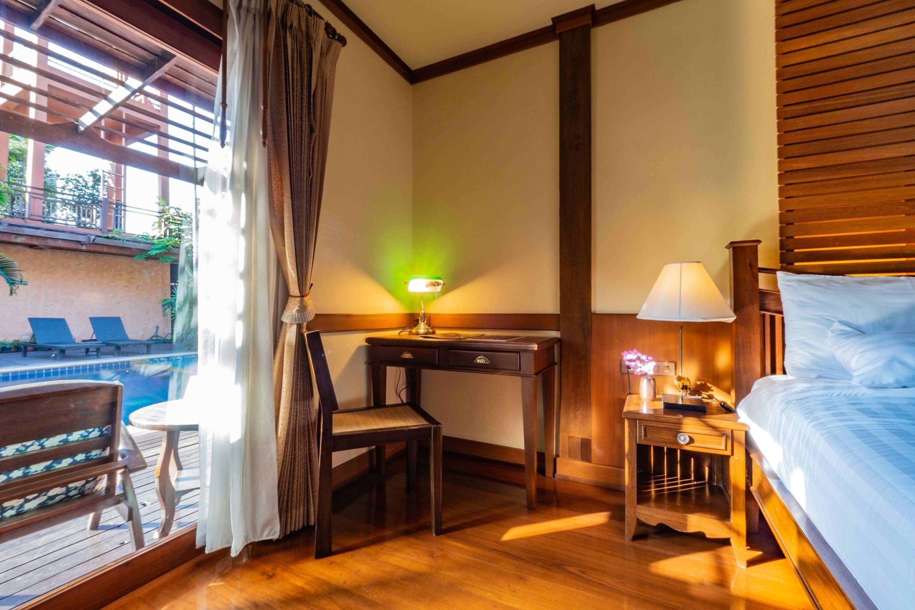 Ein Hotelzimmer mit Holzschreibtisch, einem Sessel, einem Nachttisch mit Lampe, einem Bett und Blick auf einen Außenpoolbereich.