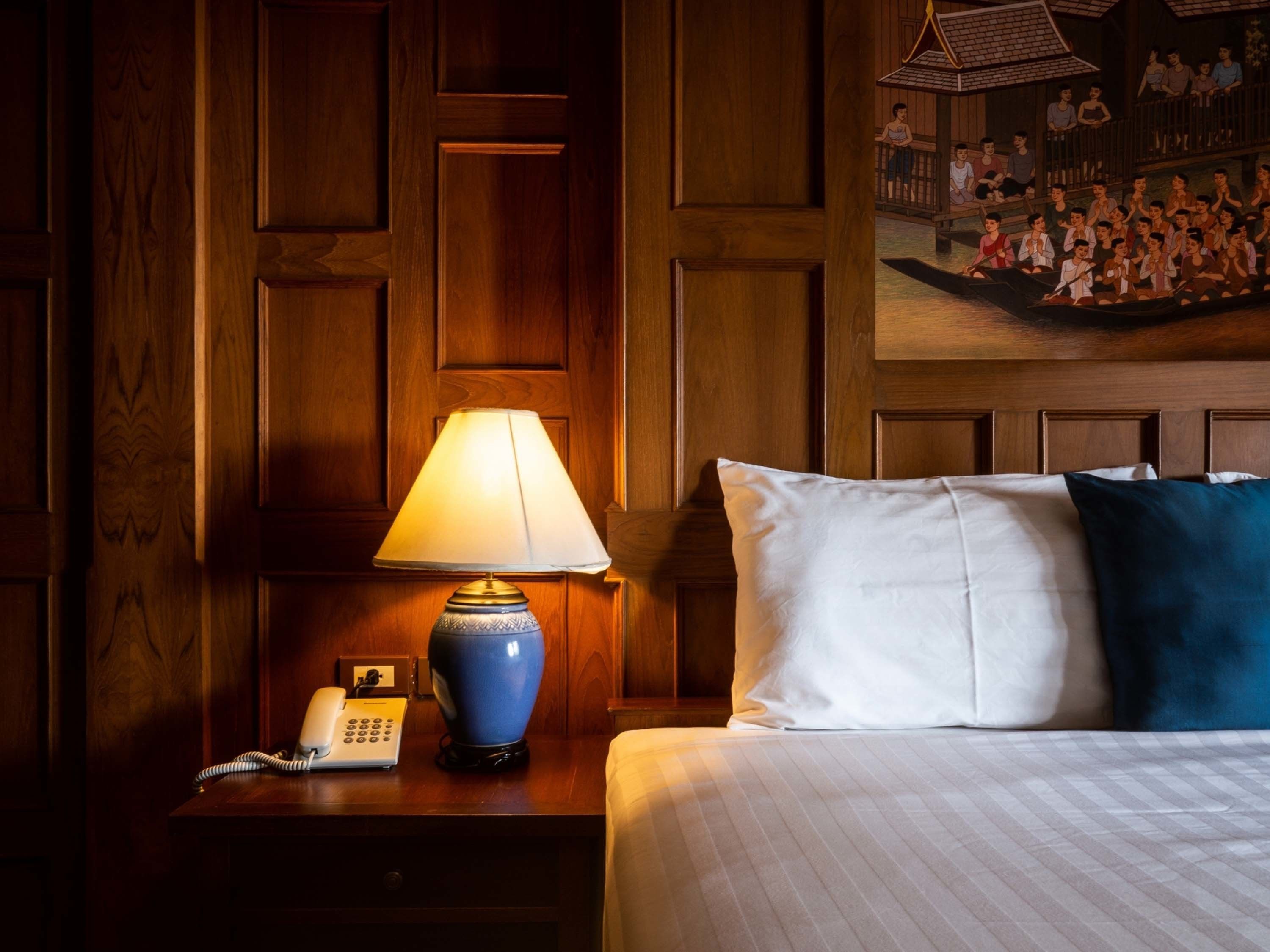 Habitación de hotel con cabecera de madera, lámpara de noche, teléfono y cama con almohadas blancas y azules.