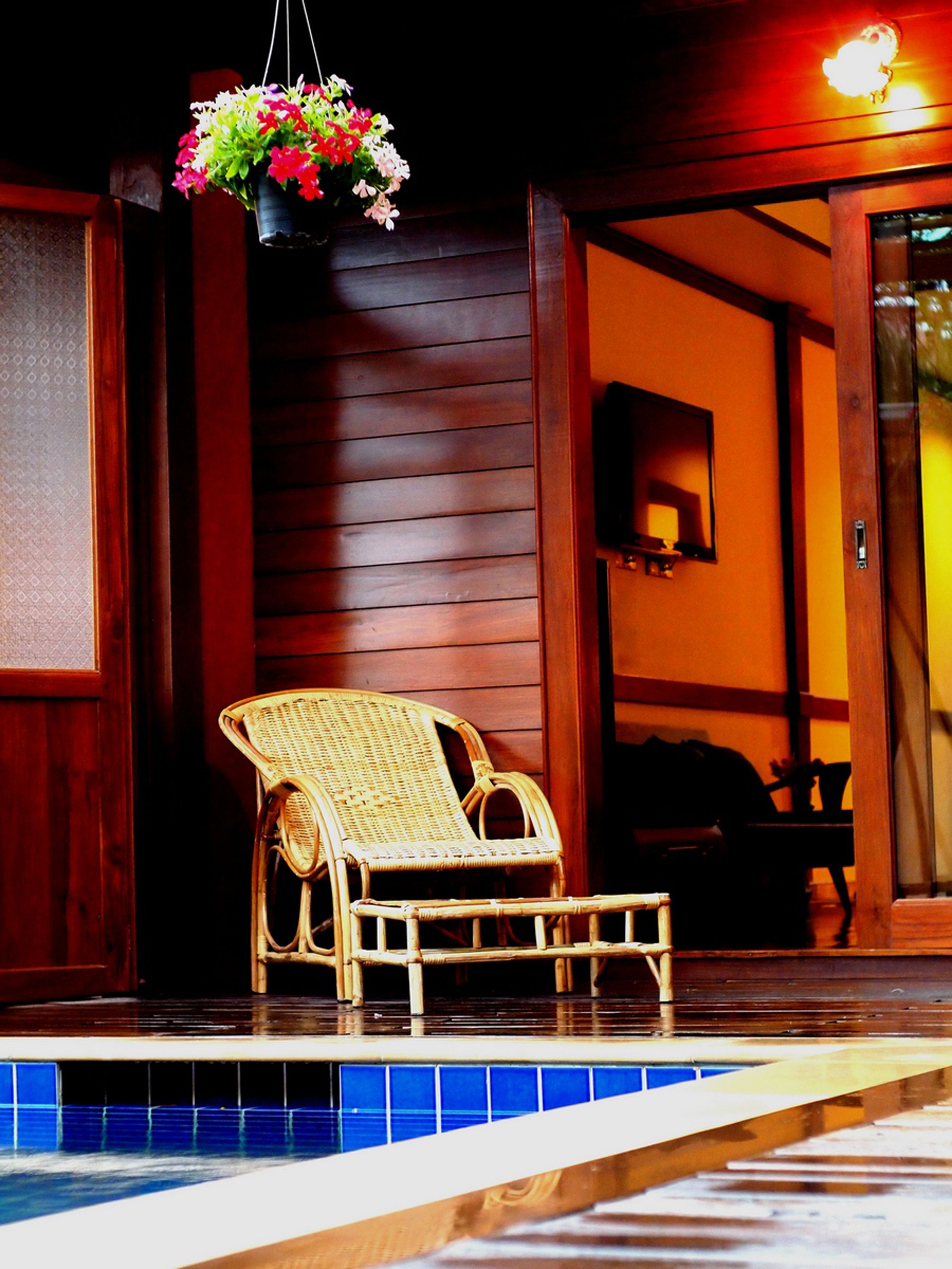 A wicker chair by a poolside, wooden walls, and a hanging flower basket.