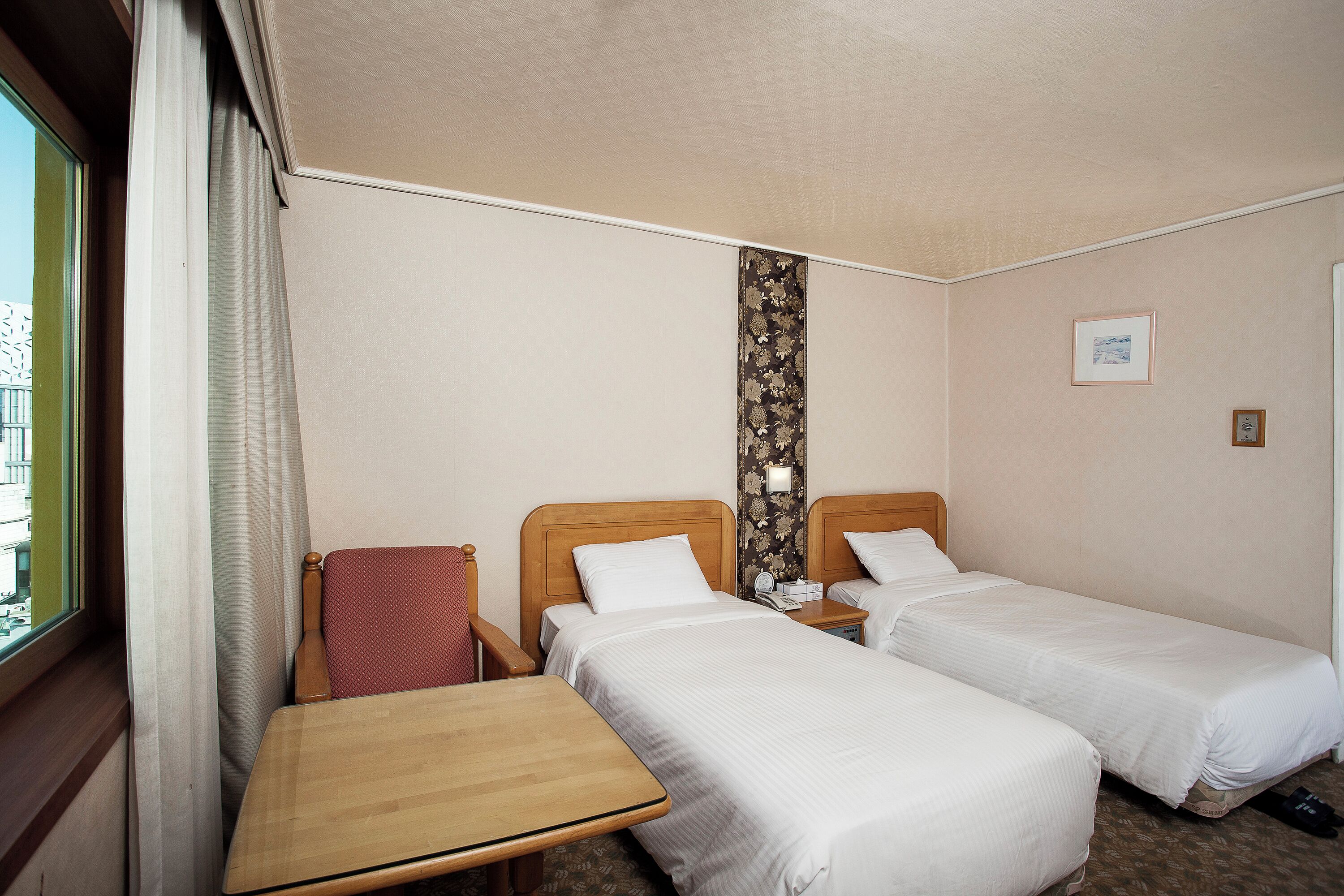 A hotel room with two single beds, a wooden table, a chair, a window with curtains, and a wall-mounted telephone.