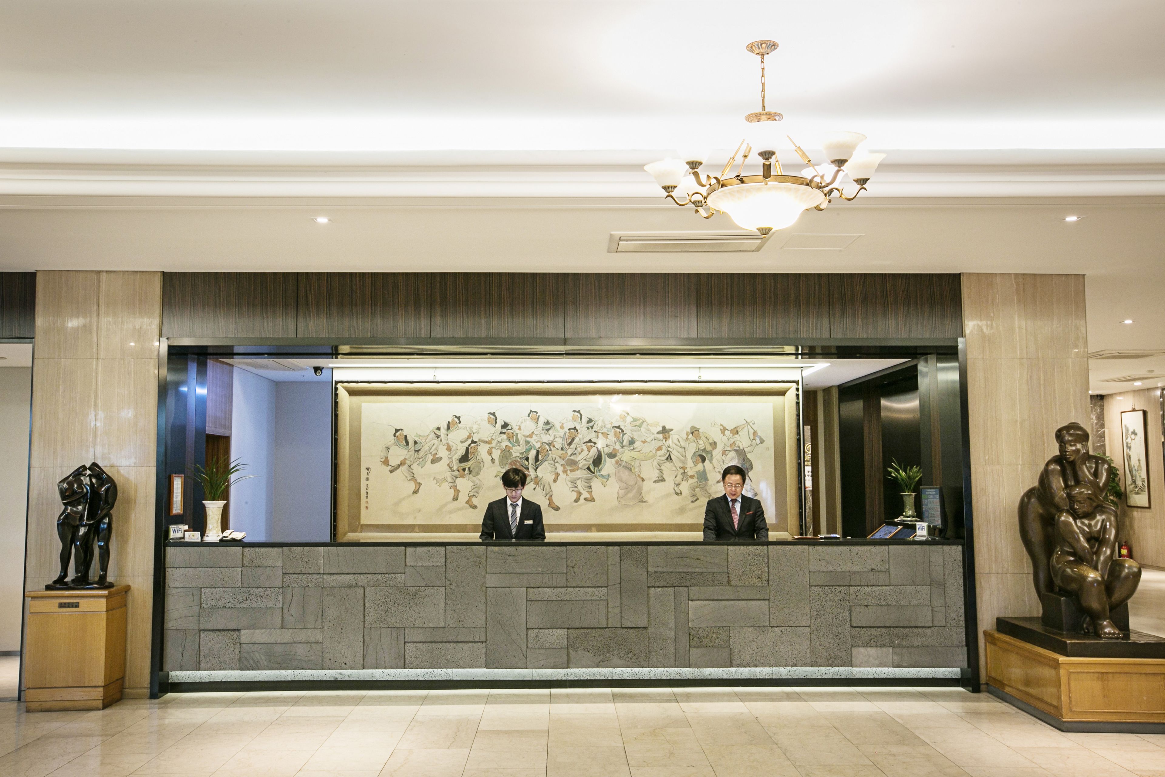 A reception desk with two staff members, a chandelier, and a large wall painting.