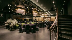 Een ruime balzaal met meerdere ronde tafels, elegante kroonluchters en een vleugelpiano.